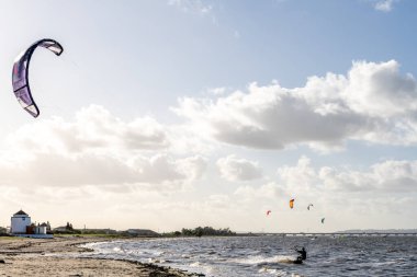 Alcochete Portekiz 19 Aralık 2025. Uçurtmacılar kumlu kıyı şeridi yakınlarında küçük binalar ve kıyı manzarası olan rüzgar enerjili tahtalara biniyorlar..