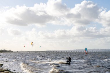 Alcochete Portekiz 19 Aralık 2025. Bir uçurtma ustası uzak köprü boyunca çoklu yelkenler görünürken dalgalı nehir suyunda manevra yapar..
