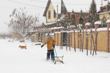 Kırmızı köpeği olan sarı ceketli bir çocuk karlı bir sokakta yürüyor. Bir çocuk kızak çekiyor. Kış eğlencesi.