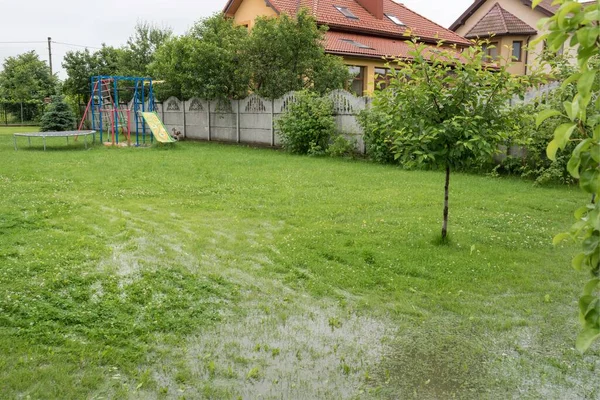 The garden and courtyard are flooded. Consequences of downpour, flood. In the background is a playground and house. Rainy summer or spring