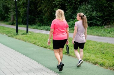 Two friends walk side by side in a lush park, enjoying their time outdoors. Both women sport varied styles and body types as they bond over daily fitness activities.