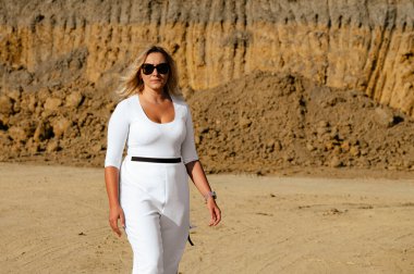 A woman wearing a white outfit strolls across a sandy construction area, with a large dirt mound in the background. Bright sunlight highlights her confident demeanor as she walks.