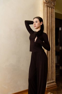 Bir kadın, ışık duvarına karşı güvenle ayakta duruyor. Koyu renk bir kıyafet giyer ve bir eli kalçasında, diğeri de başının arkasında poz verir. Ayarlar süslemeli sütunları gösterir.