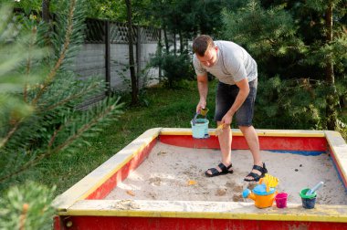 Bir baba renkli bir kum havuzunda çocuklarıyla iyi vakit geçirmekten hoşlanır. Güneşli bir günde yeşil ağaçlarla çevrili kumdan kaleler yapmalarına yardım eder..