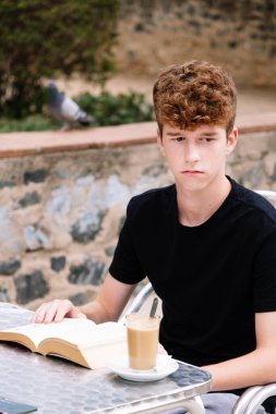 Kıvırcık saçlı genç bir çocuk barda kitap okuyor.