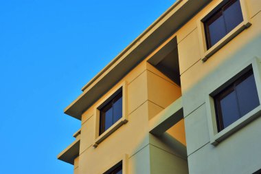 Modern bir binanın dışı, açık mavi gökyüzüne karşı alçak bir açıdan çekilmiş. Temiz çizgiler, geometrik şekiller ve yumuşak güneş ışığı çağdaş mimari tasarımı vurgular. Bu minimal ve kentsel kompozisyon emlak temaları için idealdir. 