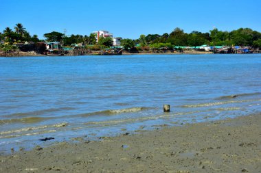 Güneşli bir günde kumlu bir kıyı şeridinde sığ nehir dalgaları yavaşça ilerler. Uzak evler ve ağaçlar karşı kıyıya uzanır. Huzurlu bir nehir kıyısında..