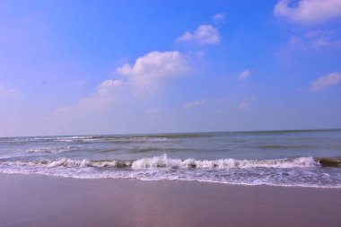 Sakin deniz manzarası, yumuşak okyanus dalgaları dağınık bulutlu açık mavi bir gökyüzünün altında kumlu bir sahile vuruyor. Gün ışığında çekilen huzurlu sahil sahnesi, deniz ufku, yumuşak dalga hareketi, ve yumuşak ıslak kum.
