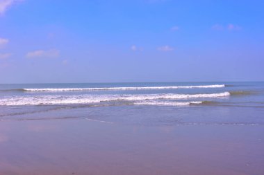 Sakin okyanus dalgaları yumuşak bulutlu açık mavi gökyüzünün altındaki kumlu bir plaja yavaşça yuvarlanır. Gün ışığında çekilen huzurlu deniz manzarası kıyı şeridini, deniz ufkunu, pürüzsüz ıslak kumu ve doğal dalga hareketini gösteriyor..