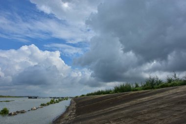 Mavi gökyüzüne karşı parlak beyaz kümülüs bulutlarının yer aldığı güzel bir sahil su yolu. Bangladeş kırsalında temiz su ve yemyeşil alan mükemmel bir yaz gününü yakalıyor..
