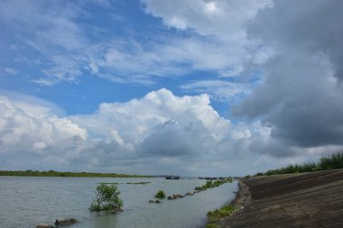 Bangladeş 'teki engin bir nehrin geniş açılı görüntüsü dramatik, bulutlu bir gökyüzü altında. Karanlık bulutlar muson mevsiminde sakin su üzerinde güçlü bir atmosfer yaratırlar..