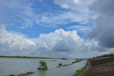 Bangladeş 'teki engin bir nehrin geniş açılı görüntüsü dramatik, bulutlu bir gökyüzü altında. Karanlık bulutlar muson mevsiminde sakin su üzerinde güçlü bir atmosfer yaratırlar..