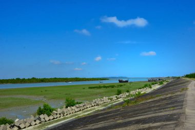 Açık mavi gökyüzünün altında alçak gelgit sırasında açık nehir yatağında dinlenen balıkçı tekneleri. Olay yeri, teknelerin gelgitte sabit kaldığı ve sular yükseldiğinde tekrar yüzeceği gelgit döngülerini belgeliyor..