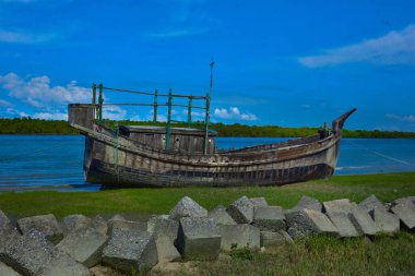 Çimenli bir nehir kıyısında dinlenen terk edilmiş eski ahşap bir tekne. Ön planda kıyı koruması için beton tetrapodlar yer alıyor. Parlak mavi gökyüzü ve uzak mangrov ağaçlarıyla huzurlu bir tropikal manzara. Geleneksel gemi.