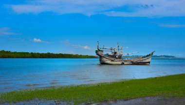 Çimenli bir nehir kıyısında dinlenen terk edilmiş eski ahşap bir tekne. Ön planda kıyı koruması için beton tetrapodlar yer alıyor. Parlak mavi gökyüzü ve uzak mangrov ağaçlarıyla huzurlu bir tropikal manzara. Geleneksel gemi.