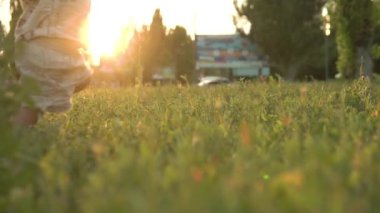 Çocuk, annesinin eşliğinde yeşil çimlerin üzerinde ilk adımlarını atıyor. Gün batımının arka planında, ağır çekimde..