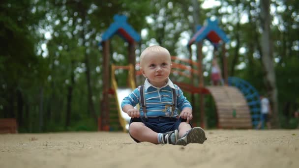 Kısa şortlu, pantalonlu, kaygısız, oyun parkında oturan ve gülümseyen tatlı sarışın bir çocuk. Mutlu çocukluk — Stok video