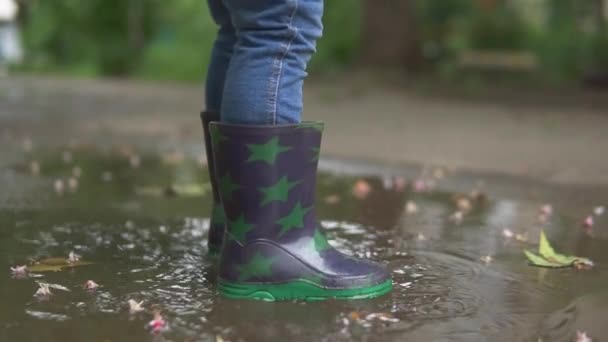 MOVIMIENTO Lento, ÁNGULO BAJO, CERRAR, DOF: Niño despreocupado con botas de lluvia grises salta al charco grande. Niño irreconocible con botas de goma nuevas salta en el charco de cristal en la acera. — Vídeo de stock