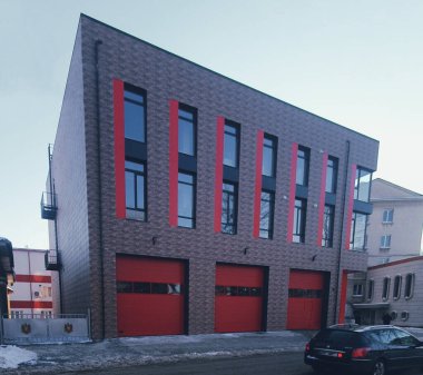 Kırmızı aksanlı ve geniş pencereli çağdaş itfaiye binası, üç garaj kapısı, modern mimari tasarımı ön plana çıkaran kentsel bir peyzaj.