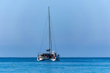 Yunanistan 'ın Lefkada kentindeki Porto Katsiki' de sakin bir sahil sahnesinde berrak bir gökyüzü altında sakin mavi sularda yüzen bir katamaran görülüyor. Huzurlu İyonya manzarası bu ünlü kumsalın doğal güzelliğini vurguluyor..
