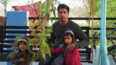 Parkta çocuklarıyla oturan genç bir Hintli. Siris 'i ya da Albizia Lebbeck' in bitki dalını tutan adam.