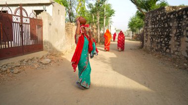 15 Nisan 2021 - Reengus, Sikar, Hindistan. Seçici odak noktası Hindu kültürlü kadın kafasına su dolu çömlek tutuyor ve kameraya bakıyor..