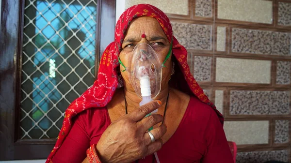 Old Indian woman infected with Covid 19 disease. Patient inhaling oxygen wearing mask with liquid Oxygen flow.