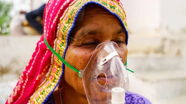 Old Indian woman infected with Covid 19 disease. Patient inhaling oxygen wearing mask with liquid Oxygen flow.