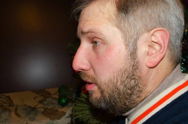 Orta yaşlı sakallı bir adam süslü Noel ağacının yanına bakıyor.