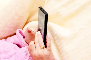 Yatakta rahatça dinlenirken akıllı telefon kullanan pembe cüppeli bir kadın.
