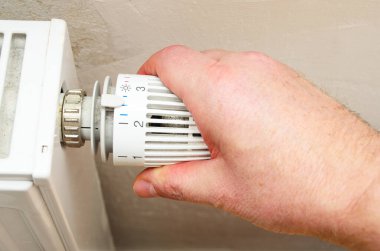 Bir adamın elinin beyaz termostatı ısıtma radyatörüne çevirmesi, artan enerji fiyatlarını, ev giderlerini ve kış mevsiminde tasarruf edilen enerjiyi sembolize ediyor  