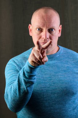 Mavi kazaklı kel bir adam elini kameraya doğrultarak doğrudan bir jest yapıyor.