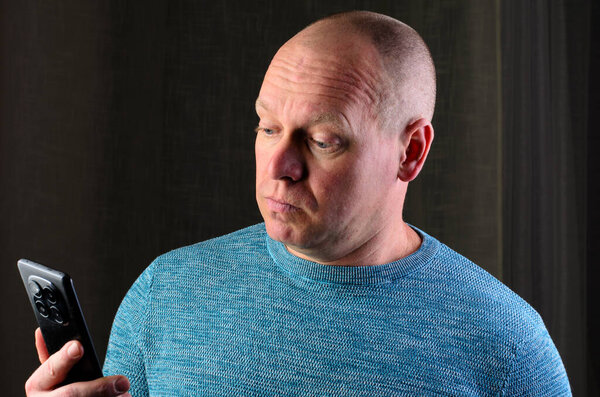 Лысый мужчина в синем свитере смотрит на свой телефон с беспокойным выражением лица, возможно читая плохие новости