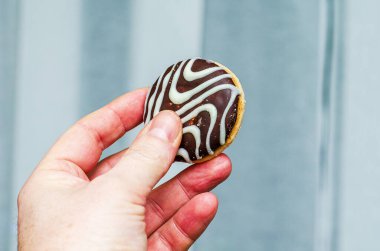 Yakın plan çekimde, siyah çikolata ve karışık beyaz krema desenleriyle kaplı yuvarlak bir bisküviyi nazikçe tutan bir insan görülüyor.