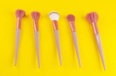 Sarı arka planda sıralanmış pembe tüylü ve leylak saplı beş makyaj fırçası koleksiyonu