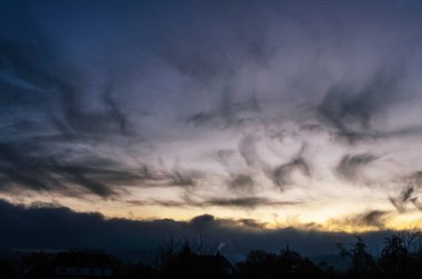 Gökyüzü büyüleyici bir karanlık, ince bulutlar ve ufuk çizgisine bakan yumuşak, sıcak bir ışık karışımı sergiliyor, siluetli çatıların üzerinde kasvetli bir atmosfer yaratıyor.