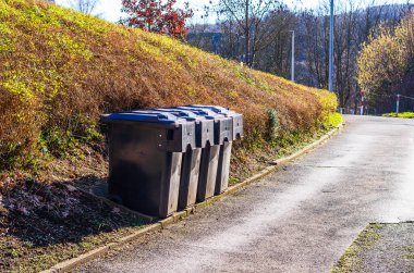 Güneşli bir günde siyah tekerlekli çöp bidonları asfalt bir yolun yanında dizilmişti.