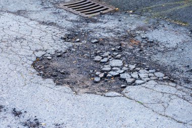 Şiddetli bir şekilde bozulmuş asfalt yol yüzeyinin yakın görüntüsü, gevşek enkazlarla dolu derin bir çukur ve çatlak kaldırımlarla çevrili, arka planda metal bir akış ızgarası var.