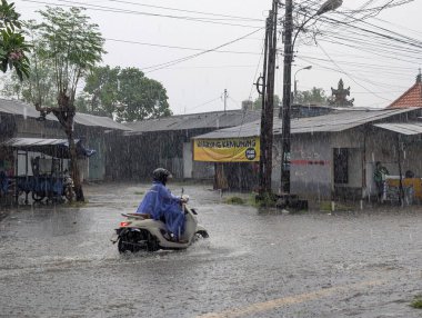 Denpasar, 19 Aralık 2025. Denpasar şehrinde sağanak yağış sırasında mavi yağmurluk giyen bir motosikletli sel basmış bir yolu geçiyor.