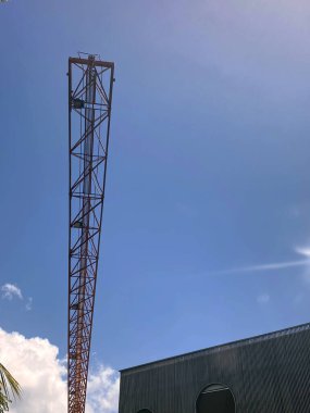 Uzun kırmızı metal kafes yapısının düşük açılı görüntüsü, muhtemelen bir iletişim direği ya da vinç, parlak mavi güneşli bir gökyüzüne karşı. Üs, modern, kıvrımlı bir binanın yanında..