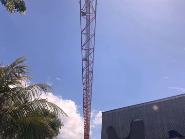 Beyaz bulutlu, parlak mavi gökyüzüne uzanan kırmızı metal bir iletişim kulesinin alçak açılı görüntüsü. Palmiye yaprakları ön planı çerçeveler, karanlık ve modern bir yapıya karşı yan yana dizilmiş..