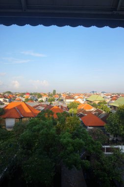 Güneydoğu Asya 'da açık mavi gökyüzü altında yemyeşil tropikal ağaçlarla çevrili geleneksel terakotta ve turuncu kiremitli çatıların yer aldığı yoğun yerleşim alanının havadan görünüşü.