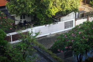 Tropikal iklimdeki yoğun bitki örtüsü olan bir yerleşim bölgesinin havadan görünüşü. Sahnede bol miktarda yeşil ağaçlar, çiçek açan bitkiler, fayanslı duvarlar ve park halindeki motosikletler yer alıyor..