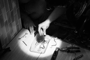 Bir tamircinin ellerini yakalayan dramatik bir siyah beyaz görüntü. Küçük makine parçaları ve aletleri, yoğun spot ışıkları altında, ahşap bir zeminde sınıflandırıyor. Tamire odaklan..