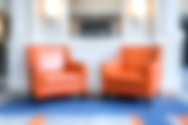 Bu resim, modern turuncu sandalyelerin şık mavi bir halı üzerinde sergilendiği, sıcak ve davetkar bir atmosfer yaratan rahat bir oturma odası sahnesini gösteriyor..