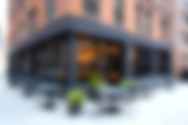 Bu görüntü, ahşap bir masa ve yeşil bir bitki süslemesinin yer aldığı rahat bir kafenin bulanık iç görüntüsünü yakalıyor ve sakin bir atmosferi çağrıştırıyor..