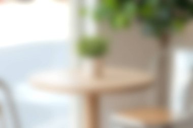 Bu görüntü, ahşap bir masa ve yeşil bir bitki süslemesinin yer aldığı rahat bir kafenin bulanık iç görüntüsünü yakalıyor ve sakin bir atmosferi çağrıştırıyor..