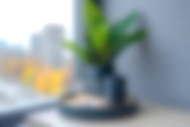 Bu bulanık görüntü, şehir manzarası penceresi olan bir masaya yerleştirilmiş kapalı bir bitkiyi gösteriyor. Sahne bir huzur ve modern yaşam hissi veriyor..