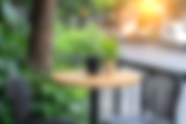 Bu görüntü, yemyeşil ve yumuşak sabah ışıklarıyla çevrili ahşap bir masa ile huzurlu bir açık kafe görüntüsü yakalar. Doğa ve yaşam tarzı temaları için ideal.