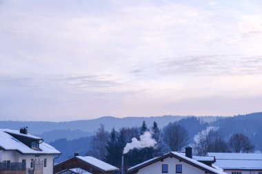 Arka planda baca dumanı ve yumuşak kış tepeleri olan karla kaplı köy çatıları. Atmosferik sabah sahnesi, kopyalama alanı. Bavyera, Almanya